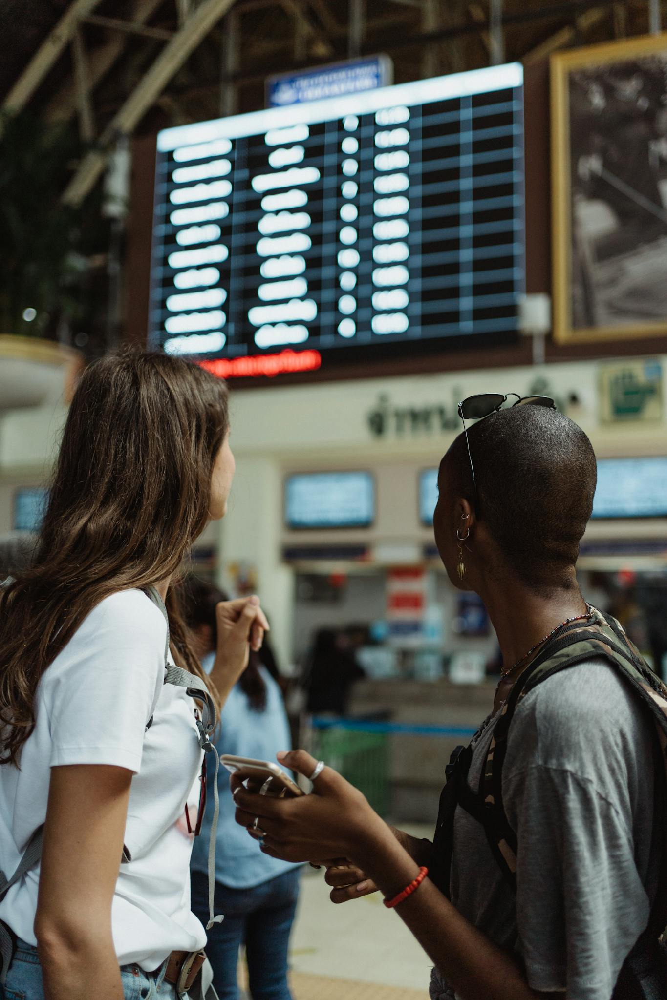 Two women checking the flight schedule on a departure board in an airport terminal, preparing for travel.